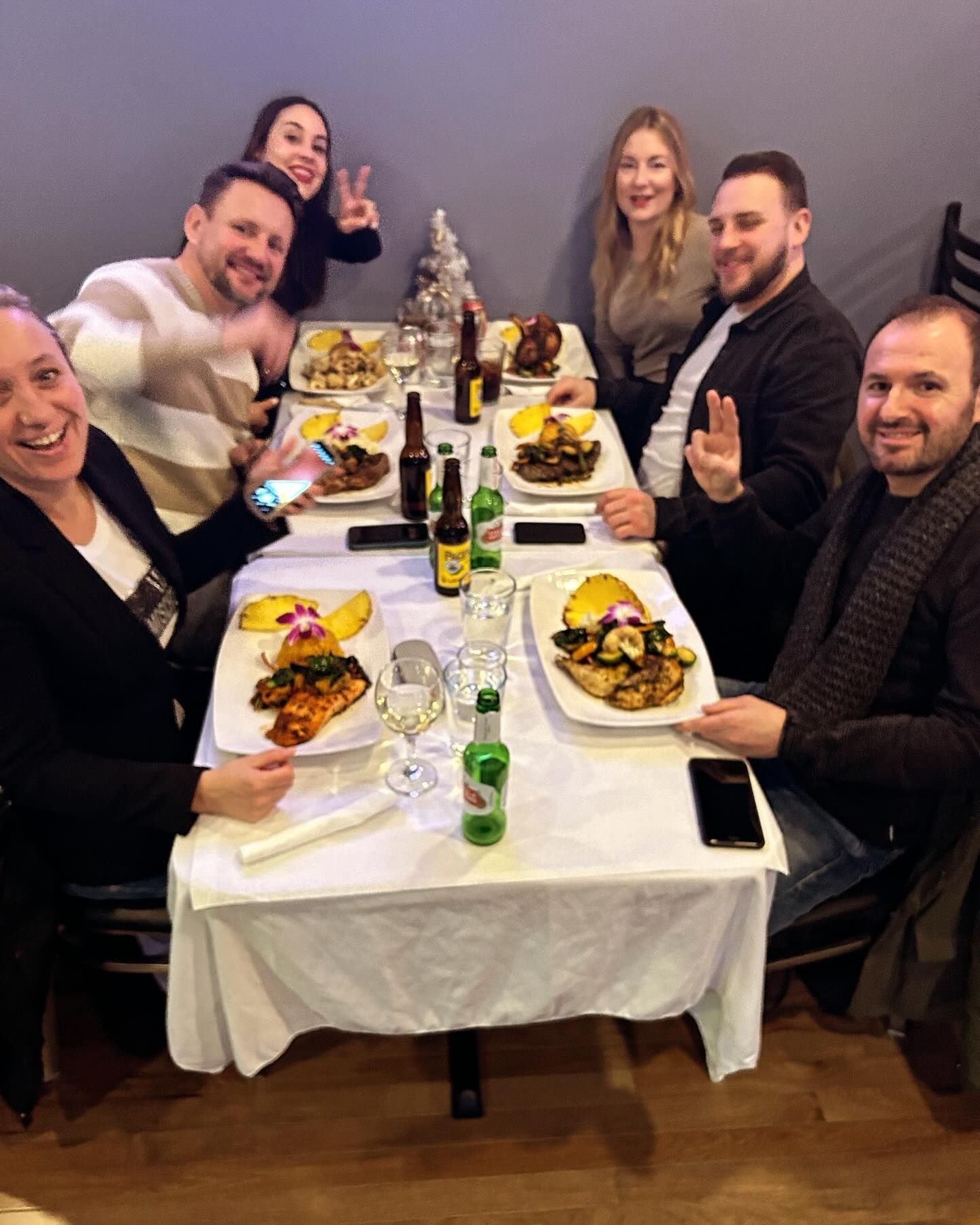 Group of seven people at a restaurant, smiling and gesturing at the camera while eating from plates on a white-covered table.