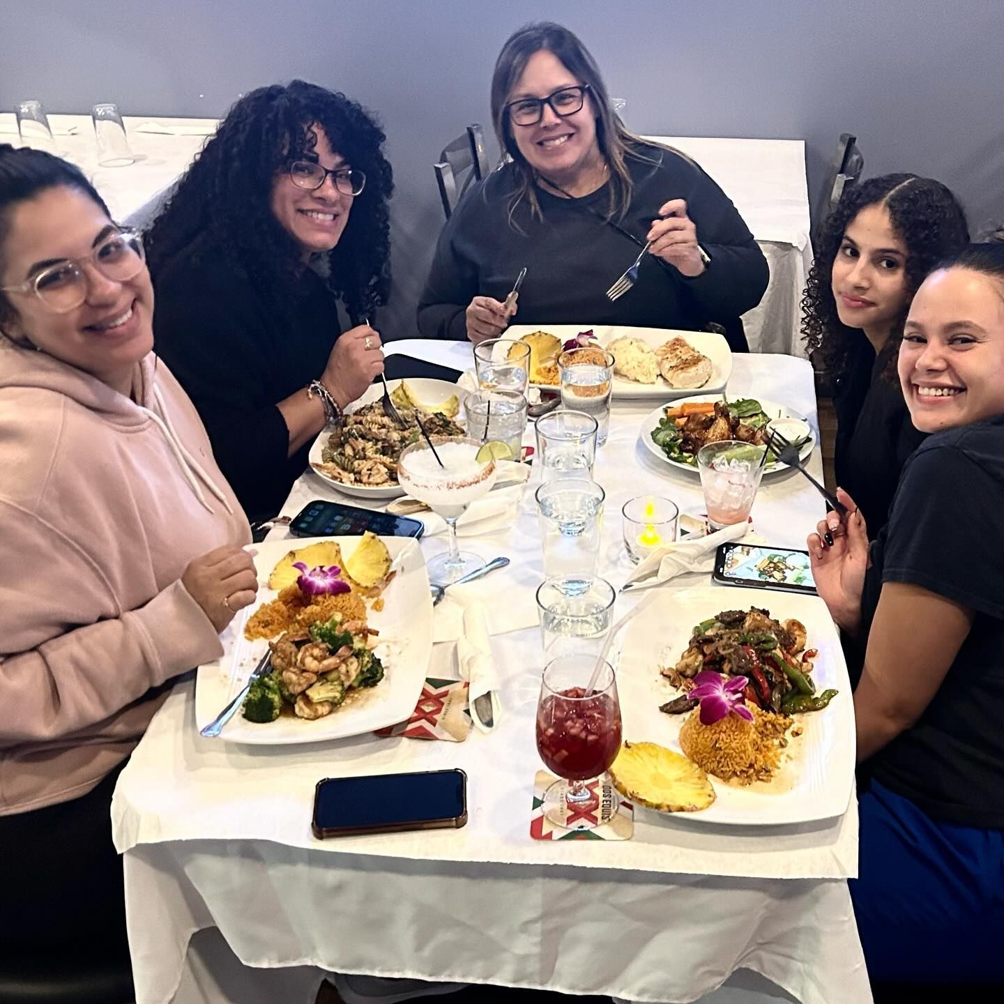 Five people at a restaurant, eating and smiling. Plates of food and drinks on the white tablecloth.