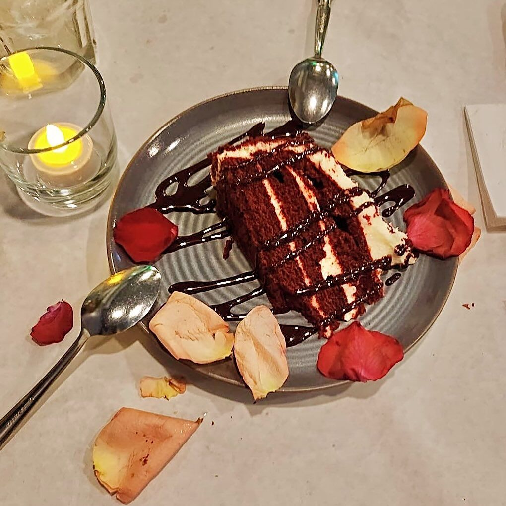 Red velvet cake slice with chocolate drizzle on a plate, rose petals, and a lit candle.
