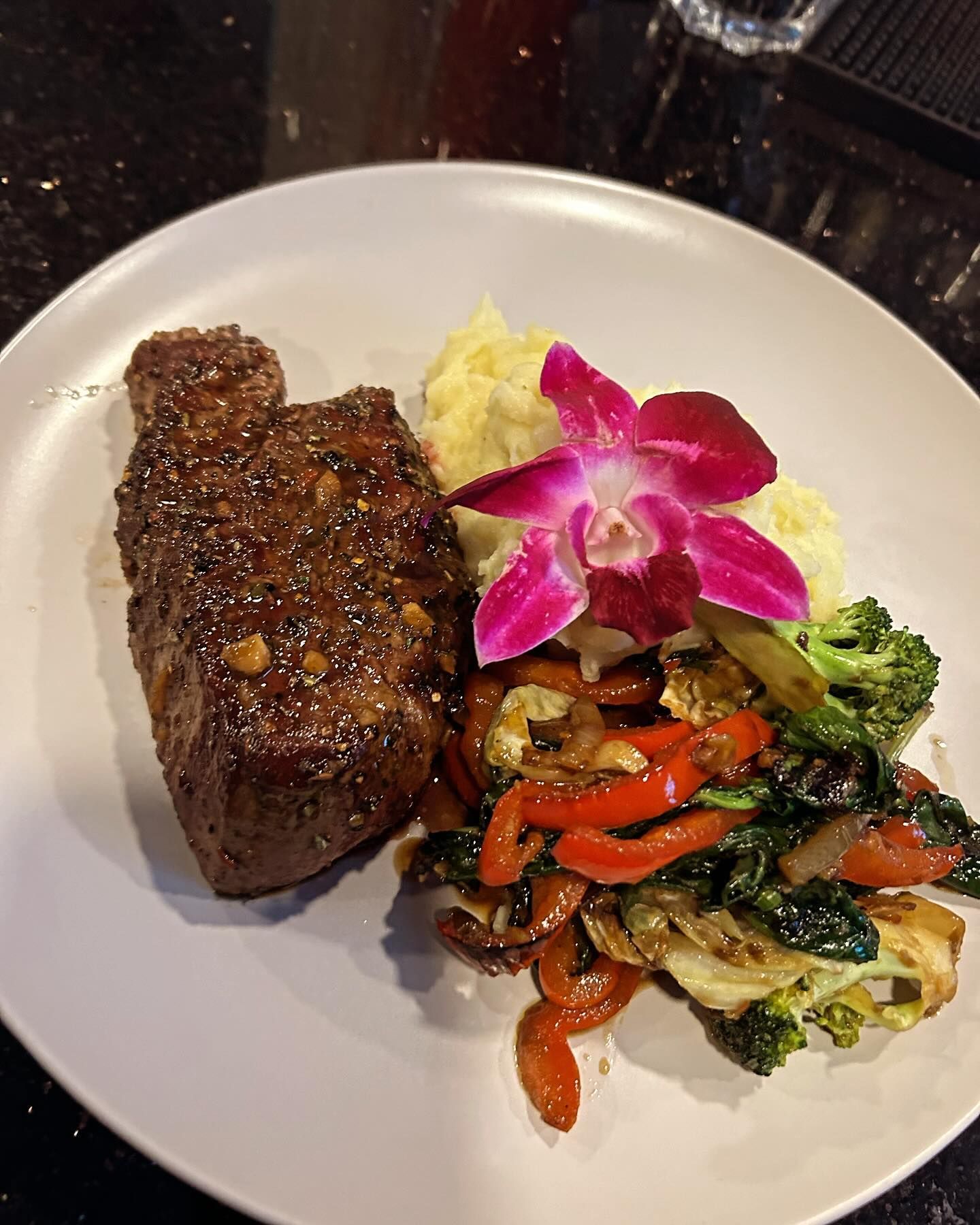 Steak with glaze, mashed potatoes, and sautéed vegetables arranged on a white plate, garnished with a flower.