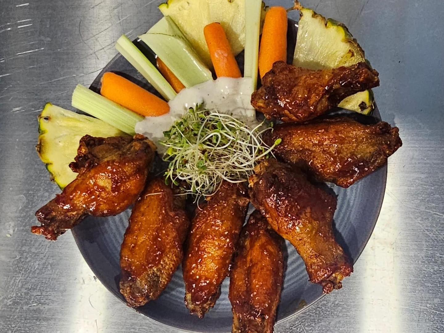 Plate of glazed chicken wings, pineapple, carrots, celery, microgreens, and dipping sauce.