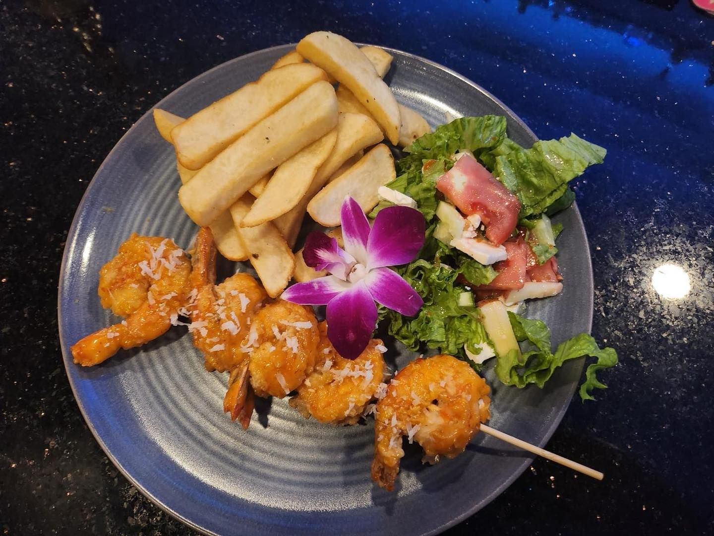 Coconut shrimp skewers with fries and salad on a blue plate, garnished with a purple orchid.