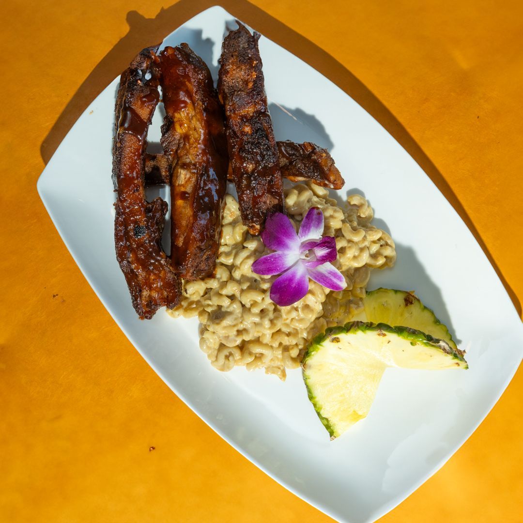 Ribs with side of grains, pineapple, and purple flower on white plate.