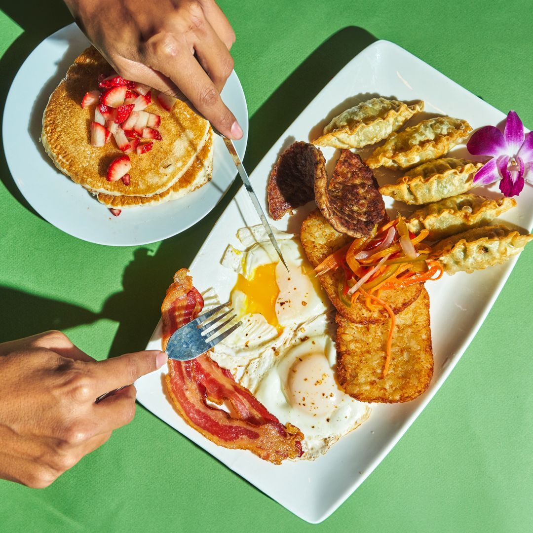 Breakfast plate: Eggs, bacon, hash brown, dumplings, sausage, and pancakes with strawberries. Two hands use a fork and knife.