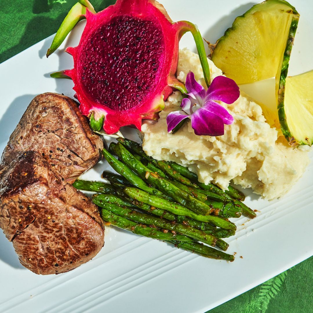 Steak, mashed potatoes, green beans, pineapple, and dragon fruit on a white plate.