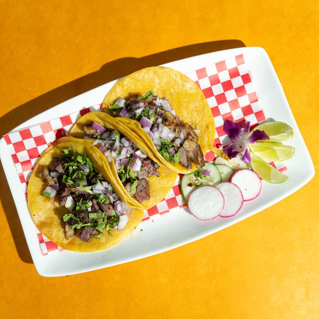 Three tacos on a white plate with red checkered paper. Garnished with lime, radish, and onion.