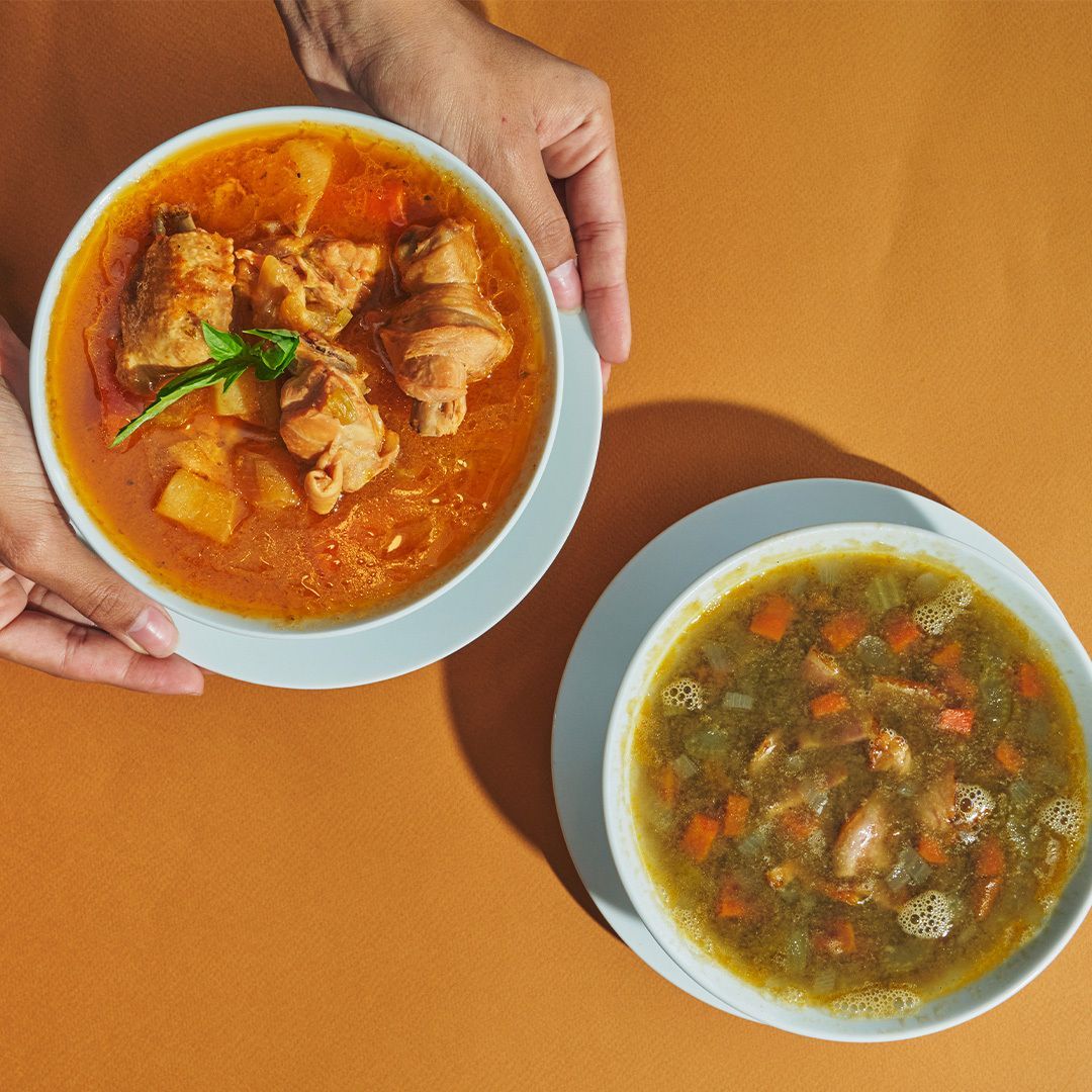 Two bowls of soup on a table; one held, one on the table. Chicken and vegetables in the soup.
