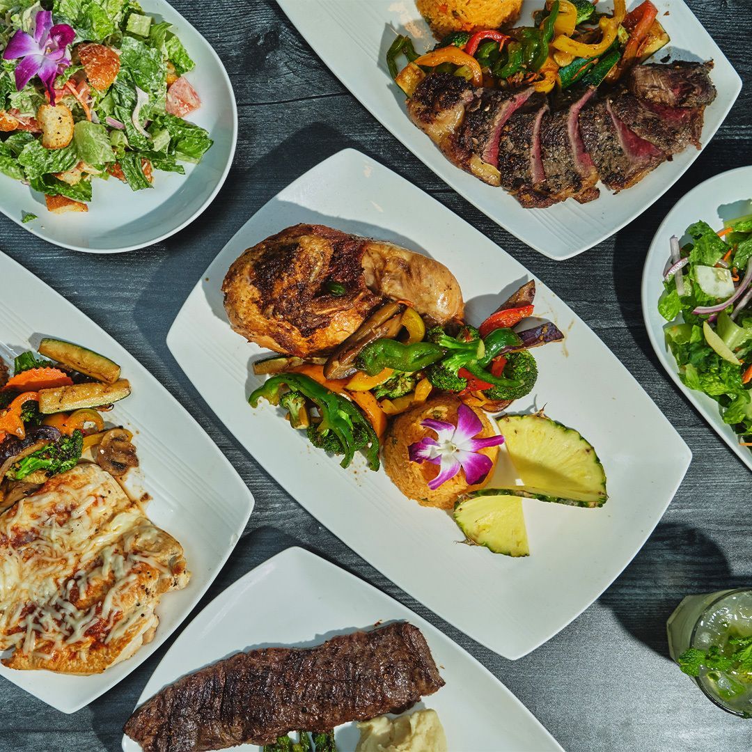 Several dishes on white plates: grilled chicken, steak, salad, and vegetables on a dark surface.