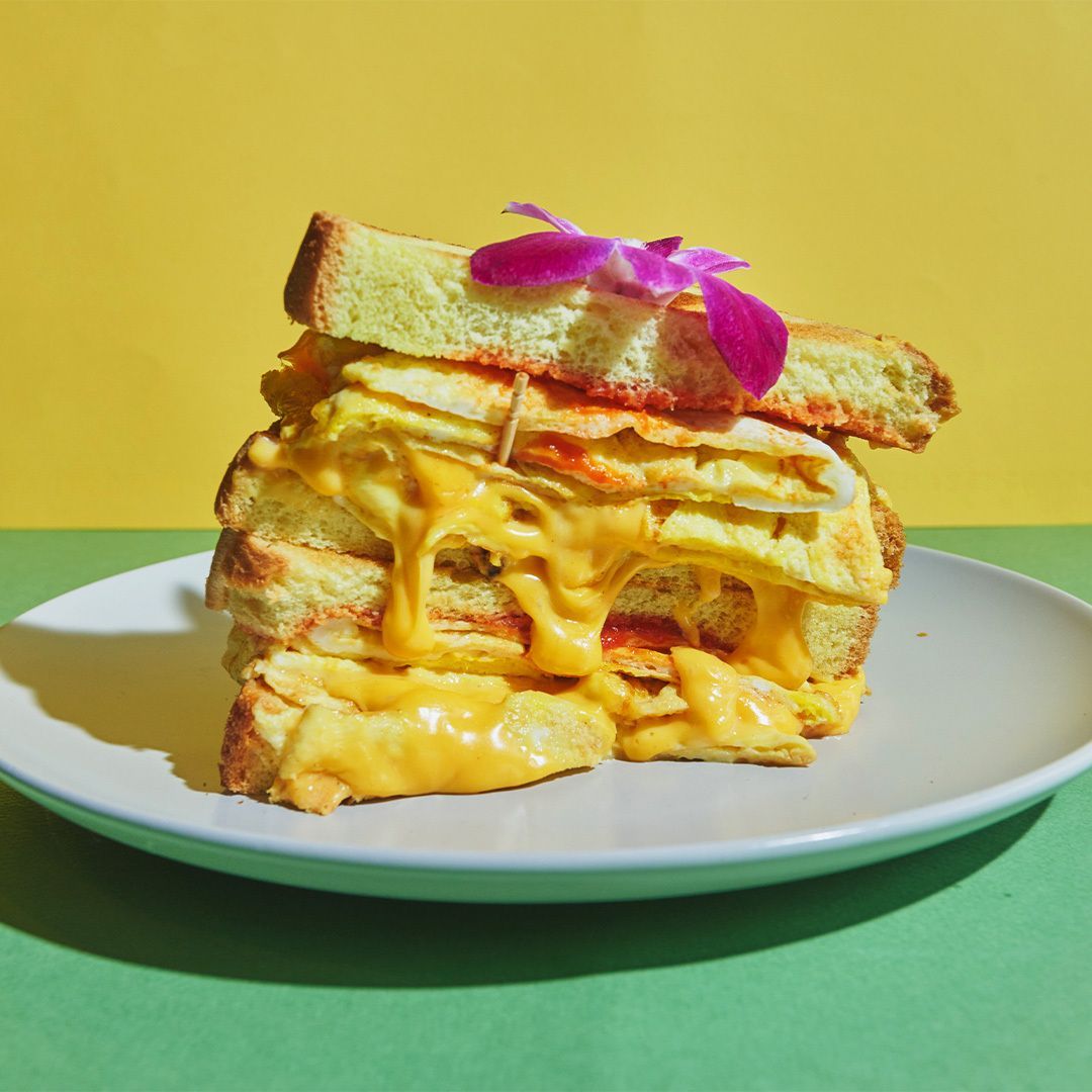 A towering sandwich with layers of cheese and egg on toasted bread, garnished with a purple flower, on a plate.