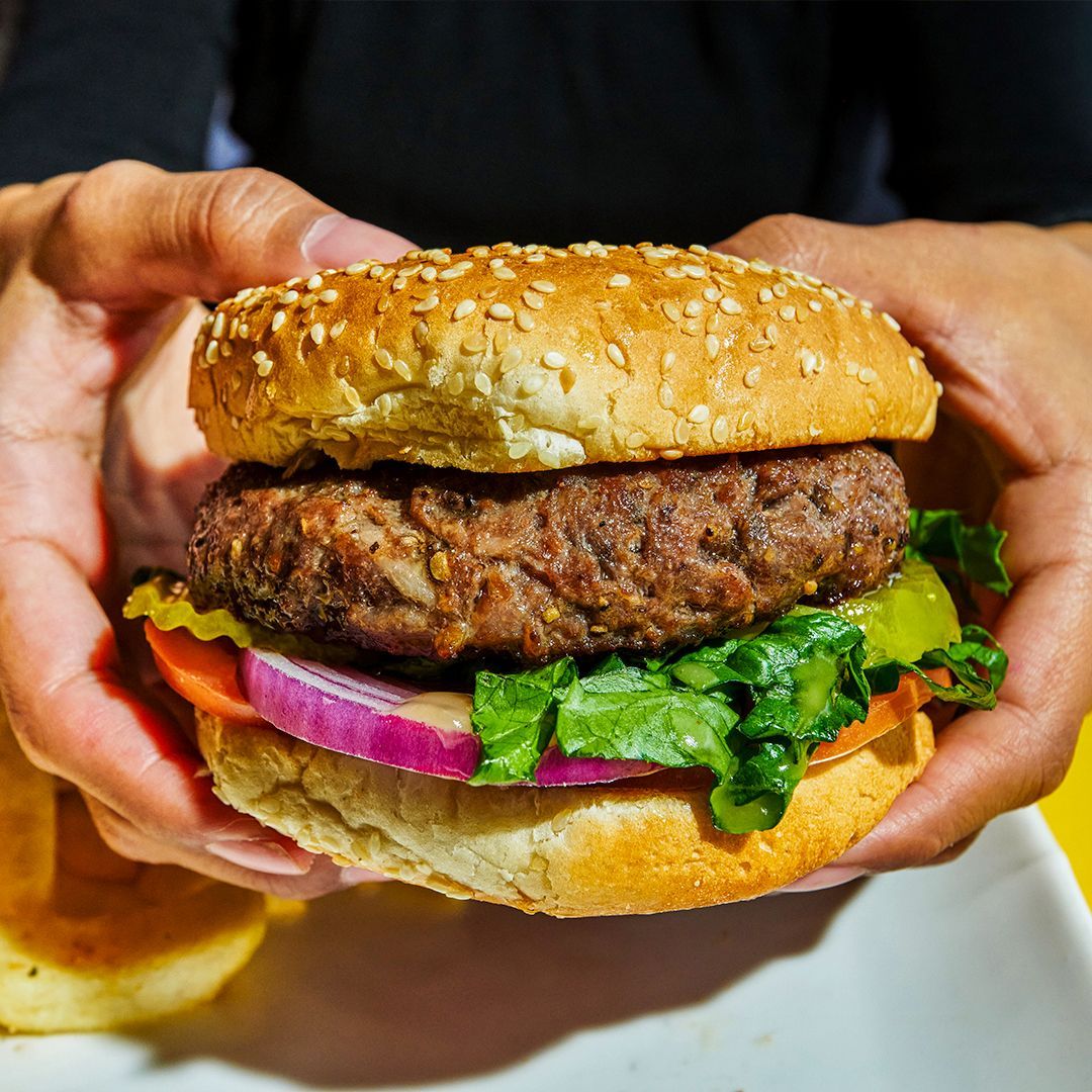 Person holding a sesame seed bun burger with patty, lettuce, tomato, and red onion.