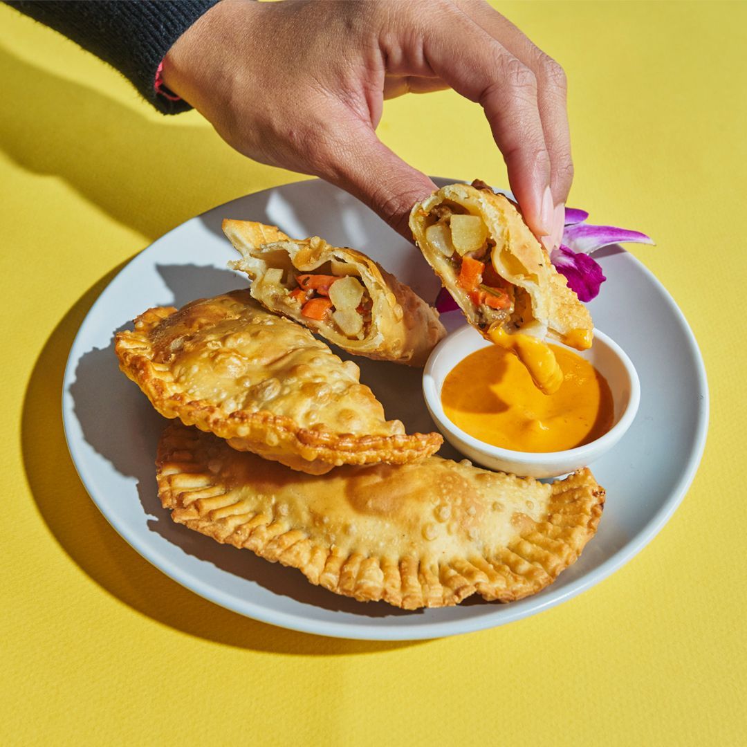 Hand holding open empanada, showing vegetable filling, next to other empanadas and sauce on a plate.