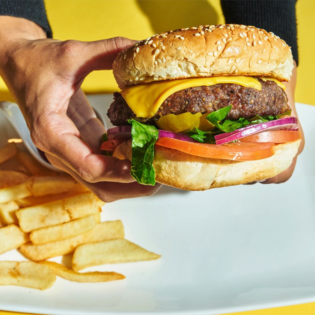 Person holding a cheeseburger with fries on a white plate; yellow background.