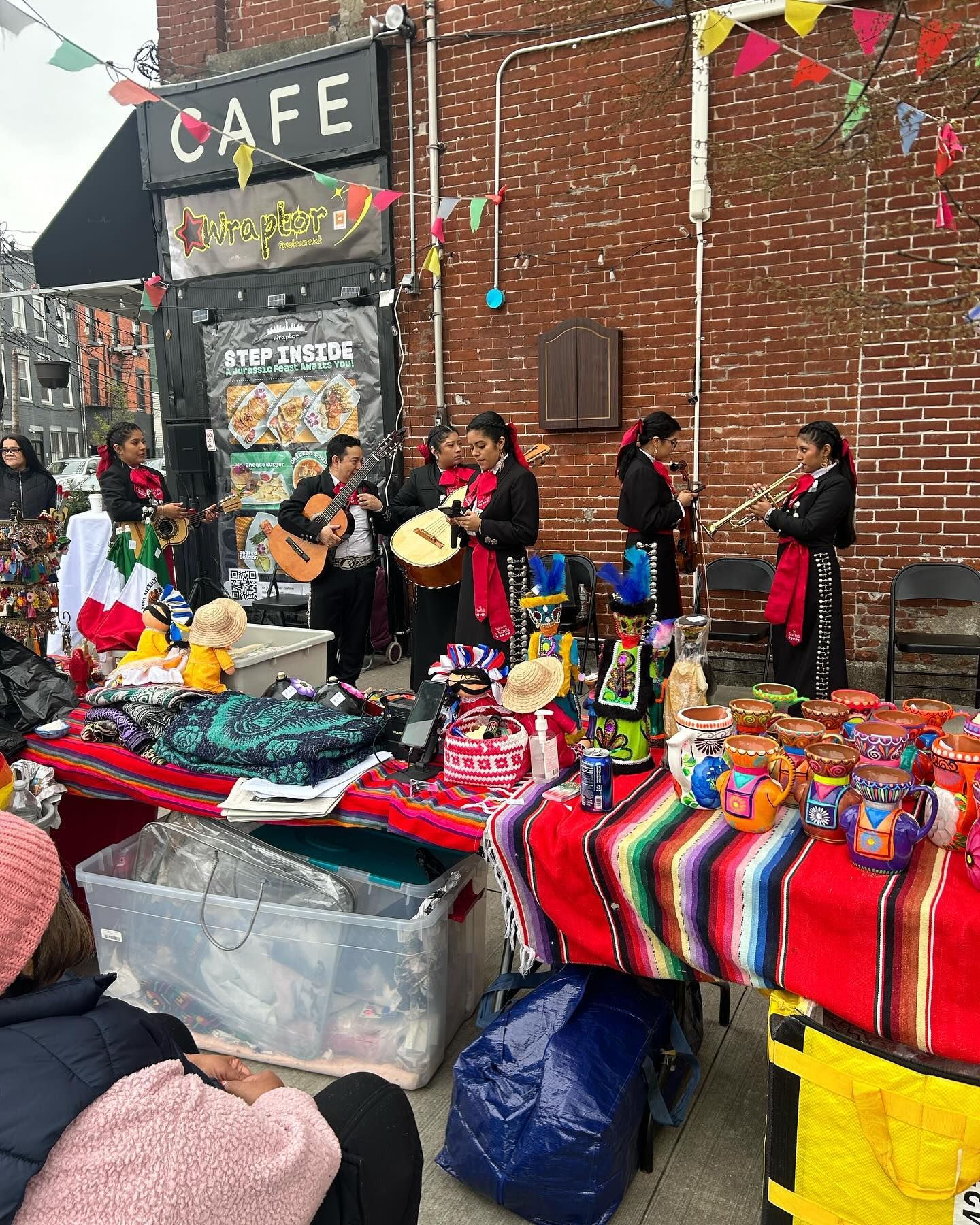 Mariachi band performs near a cafe and market stall with Mexican goods. Colorful bunting, red brick wall.