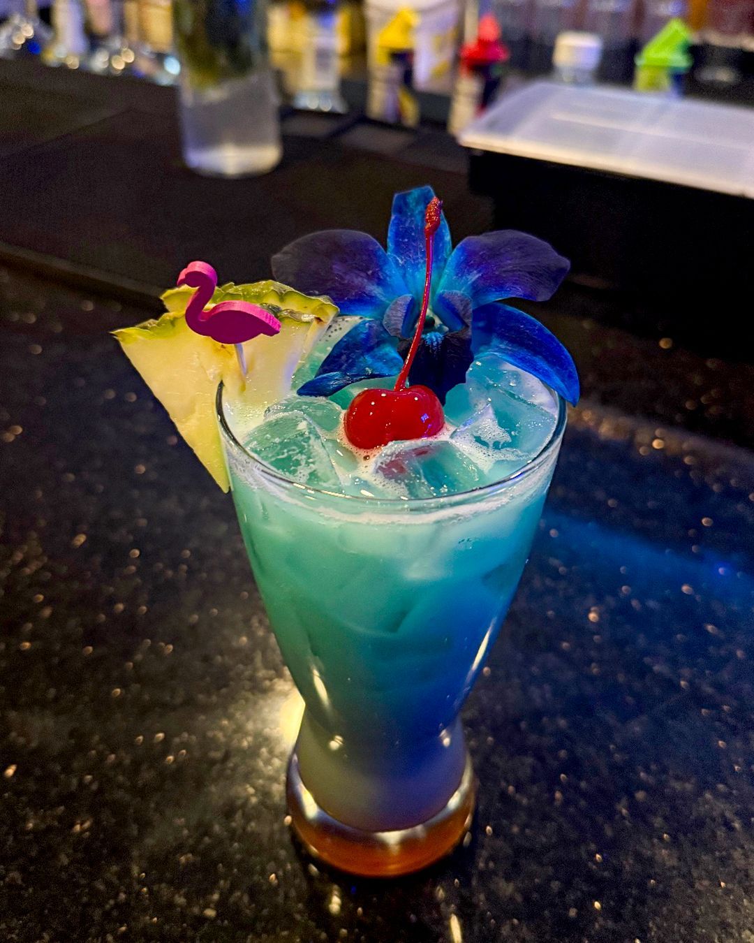 Blue cocktail with pineapple, flower, and cherry garnish, on a dark bar.