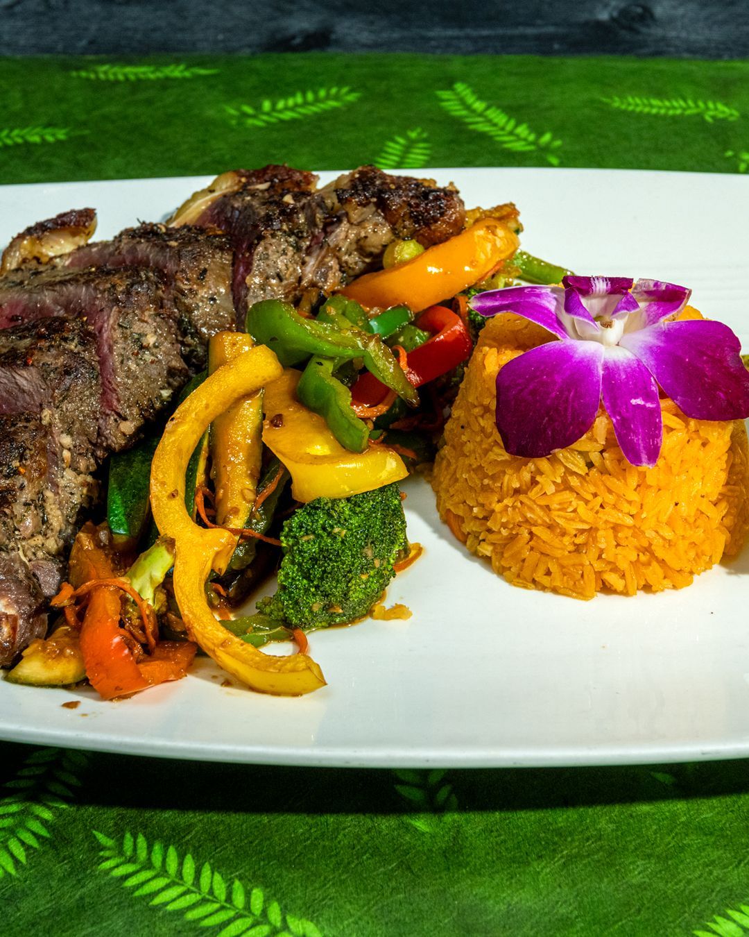 Steak, colorful sauteed vegetables, orange rice, and an orchid garnish on a white plate.