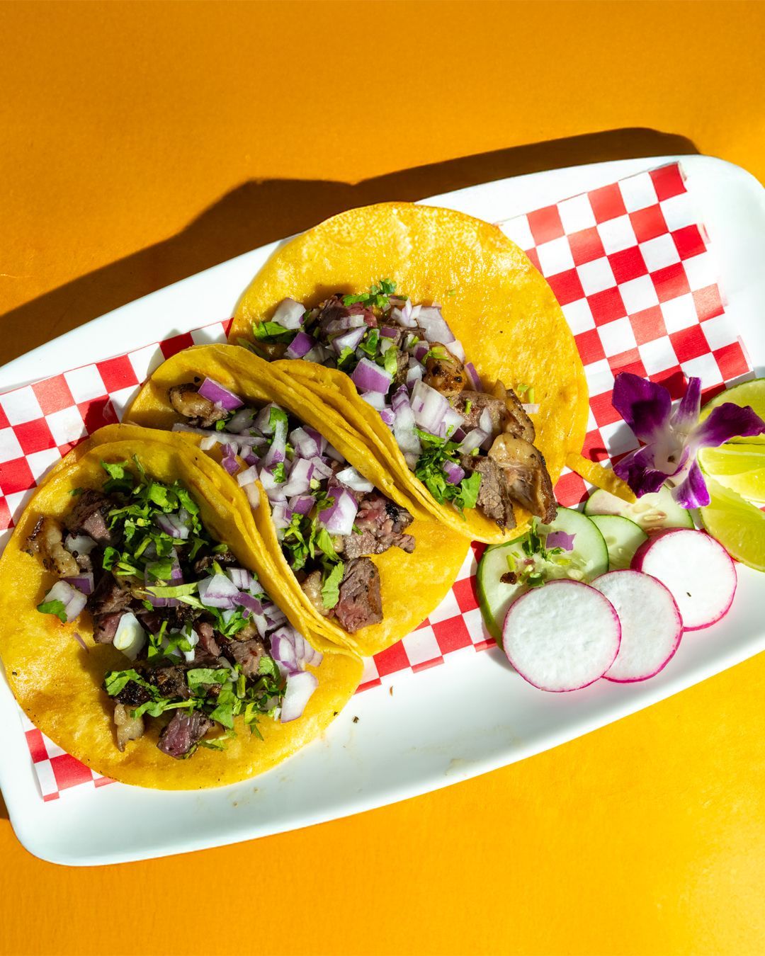 Three tacos on a plate with toppings, radishes, and lime on a red and white checkered paper.
