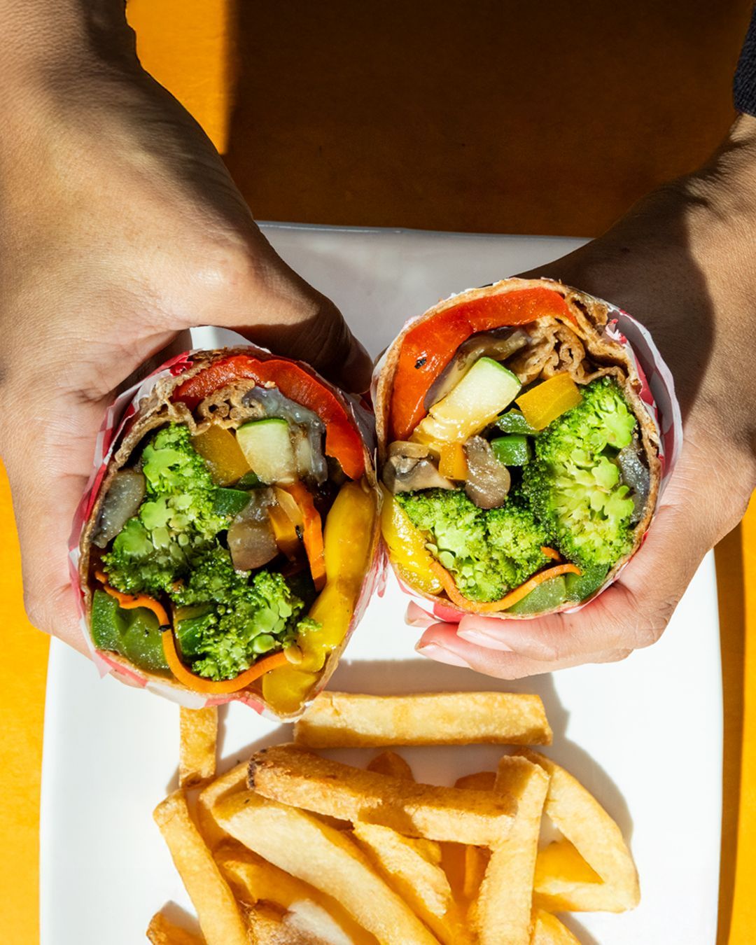 Hands holding two halves of a veggie wrap with fries on a white plate. Brightly colored vegetables are visible.