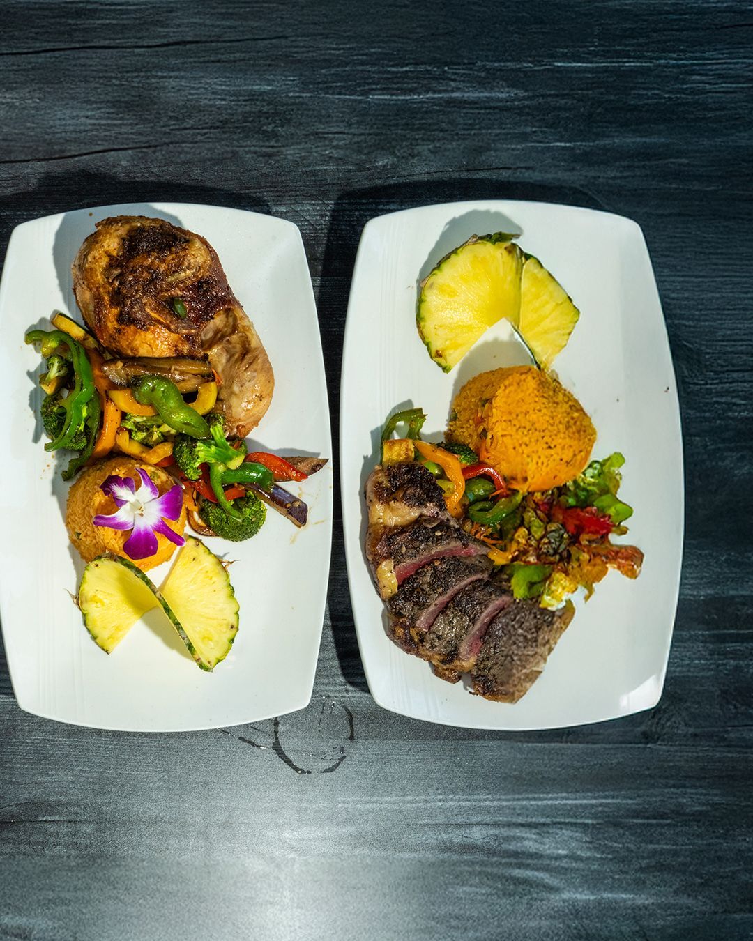 Two plates of food, one with grilled chicken and vegetables, the other with steak, pineapple, and sweet potato.