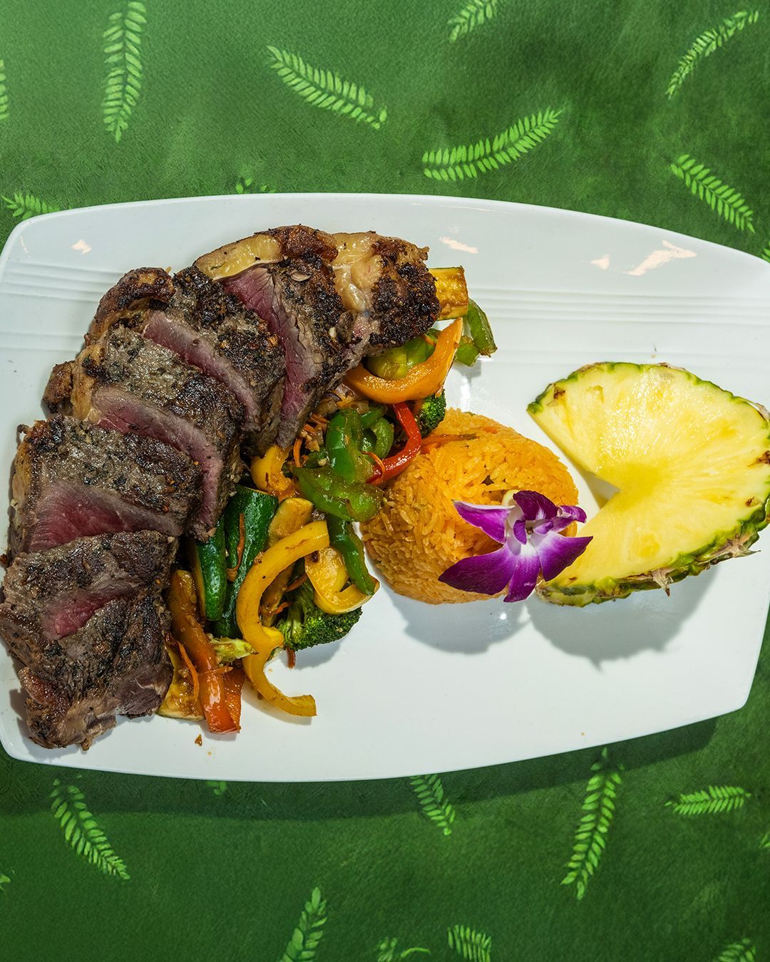 Grilled steak with colorful peppers, rice, pineapple, and a flower on a white plate.