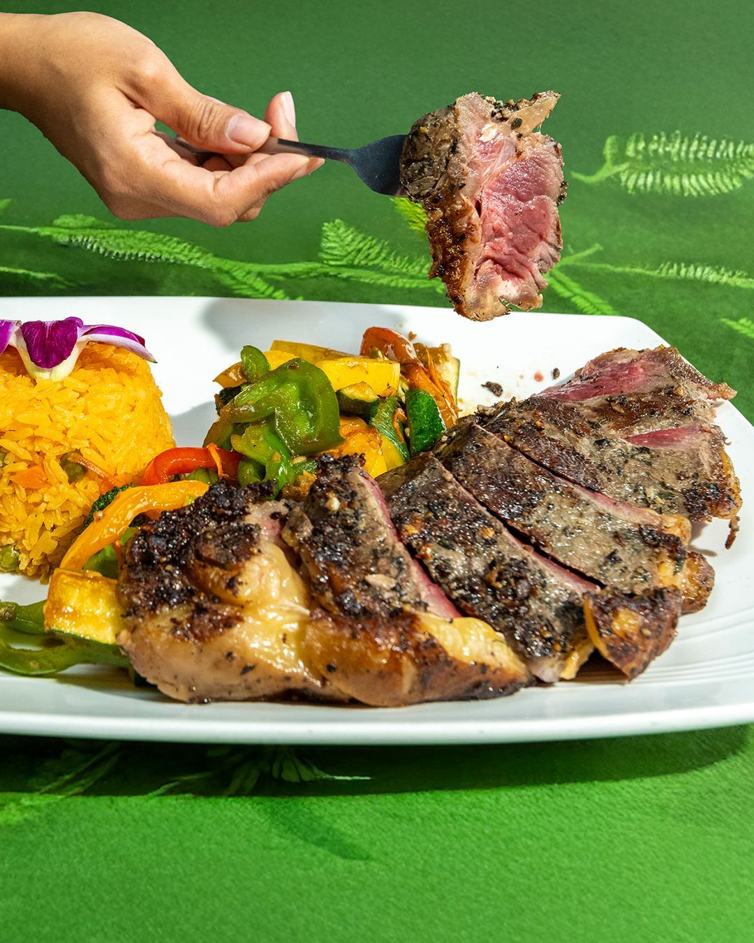 Steak on a white plate with rice and vegetables; hand holding a fork with a piece of steak.