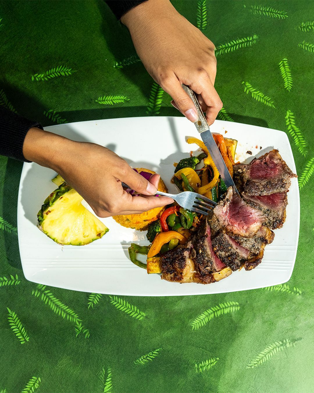 Person cutting a steak on a plate with vegetables and pineapple; green background.