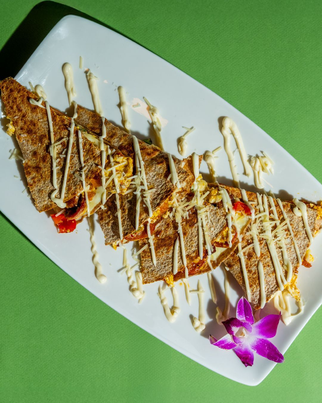Savory crepe on white plate, drizzled with white sauce, garnished with a purple flower, on green background.