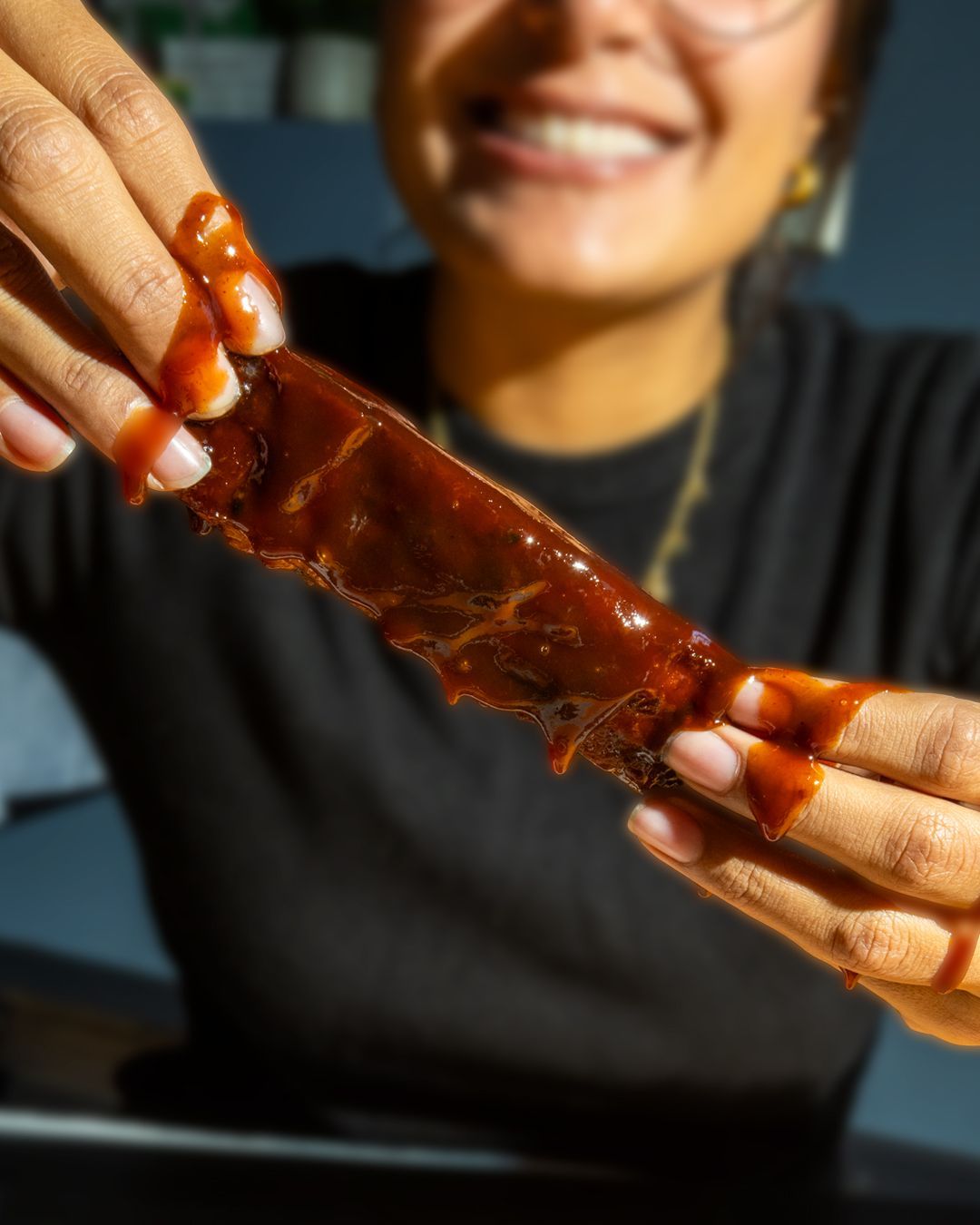 Woman holding up a saucy rib, smiling. Dark shirt, fingers covered in sauce.