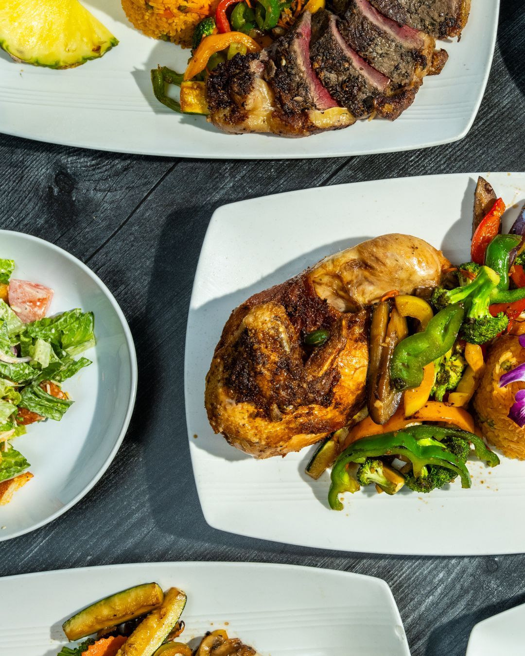 Plates of food, including steak, chicken, vegetables, and salad, on a dark wooden table.