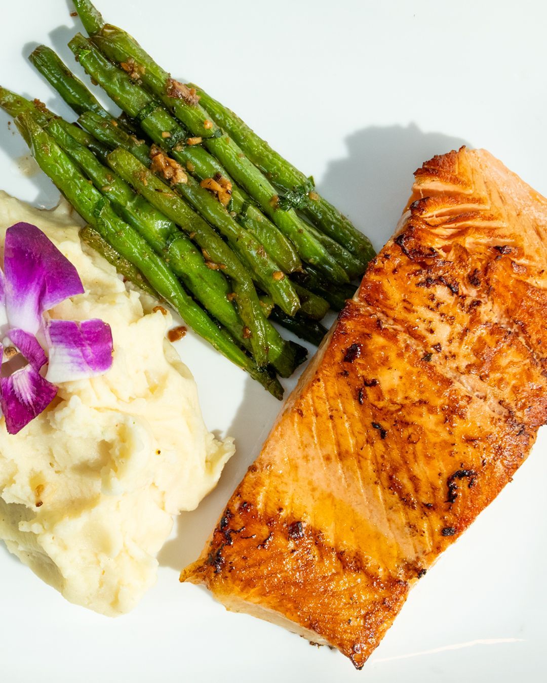 Grilled salmon with mashed potatoes and green beans on a white plate, garnished with a purple flower.