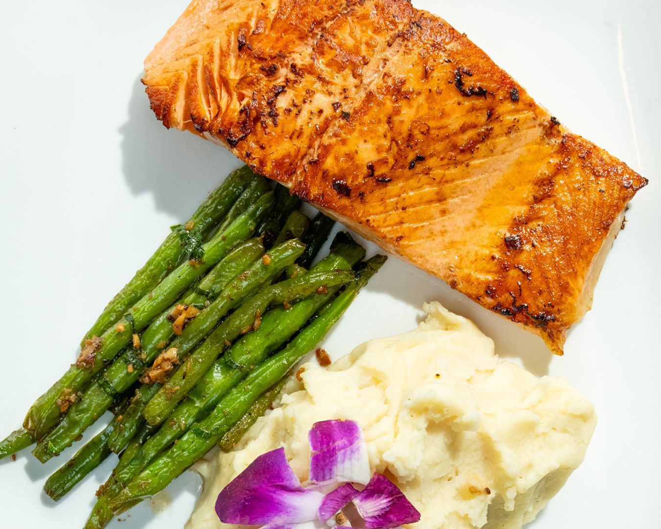 Seared salmon with green beans and mashed potatoes, garnished with a purple flower on a white plate.