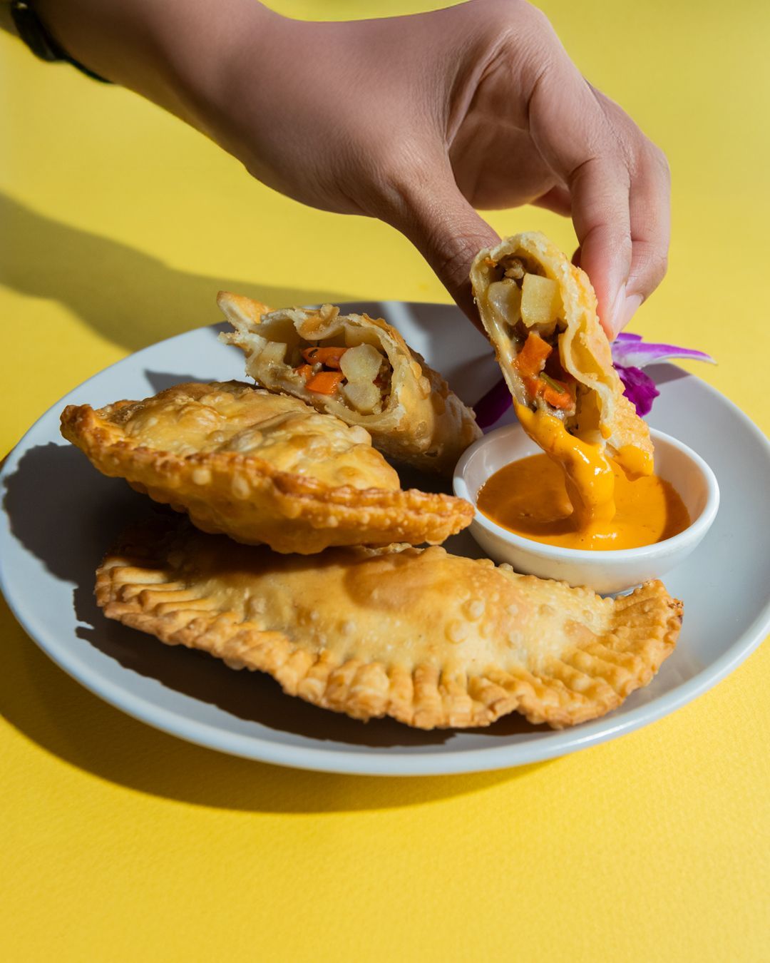 Hand dipping a golden fried pastry, filled with vegetables, into a dipping sauce on a plate. Yellow background.