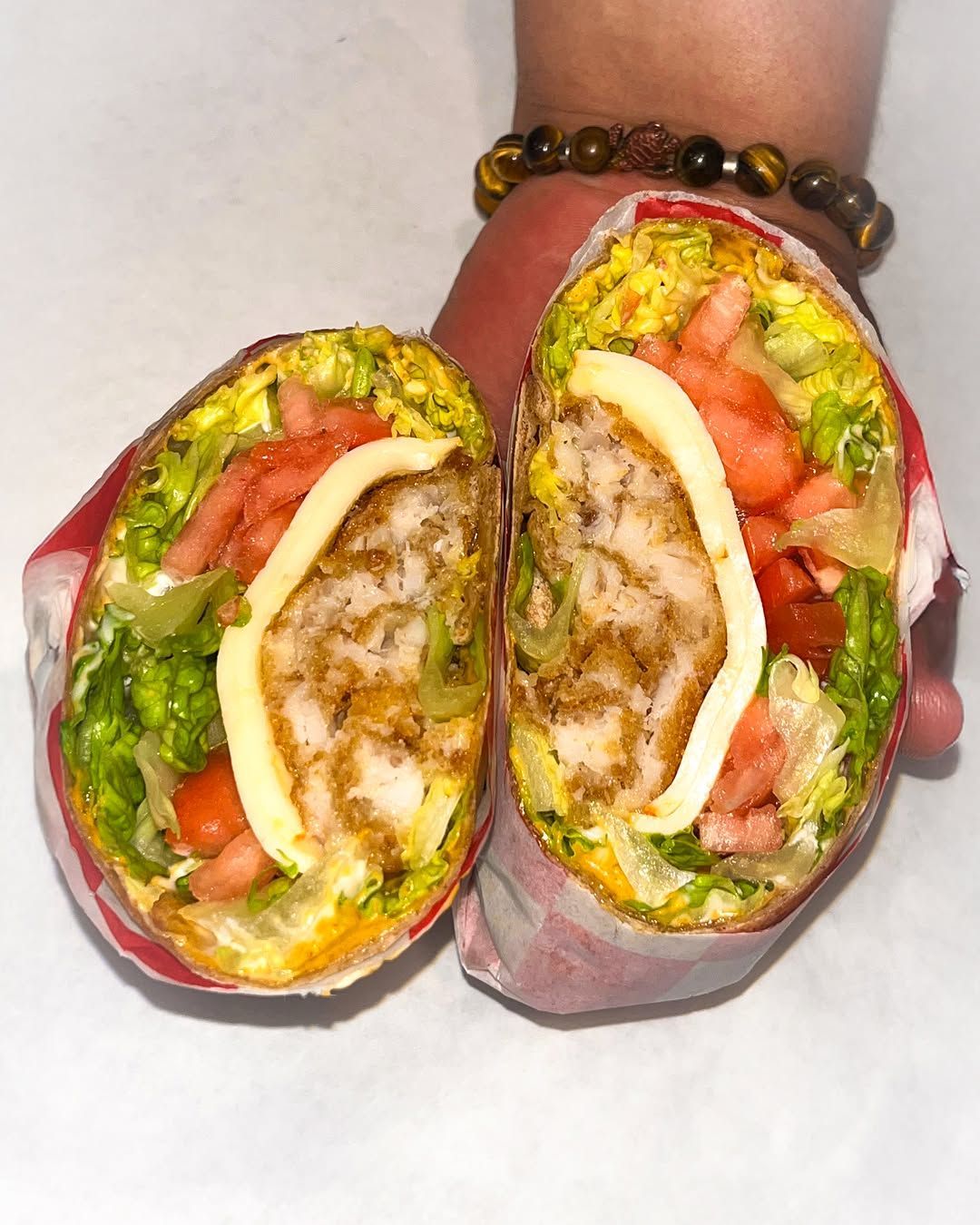 Chicken sandwich, cut in half, wrapped in paper.  Visible toppings: lettuce, tomato, cheese, dressing. Held by a hand.