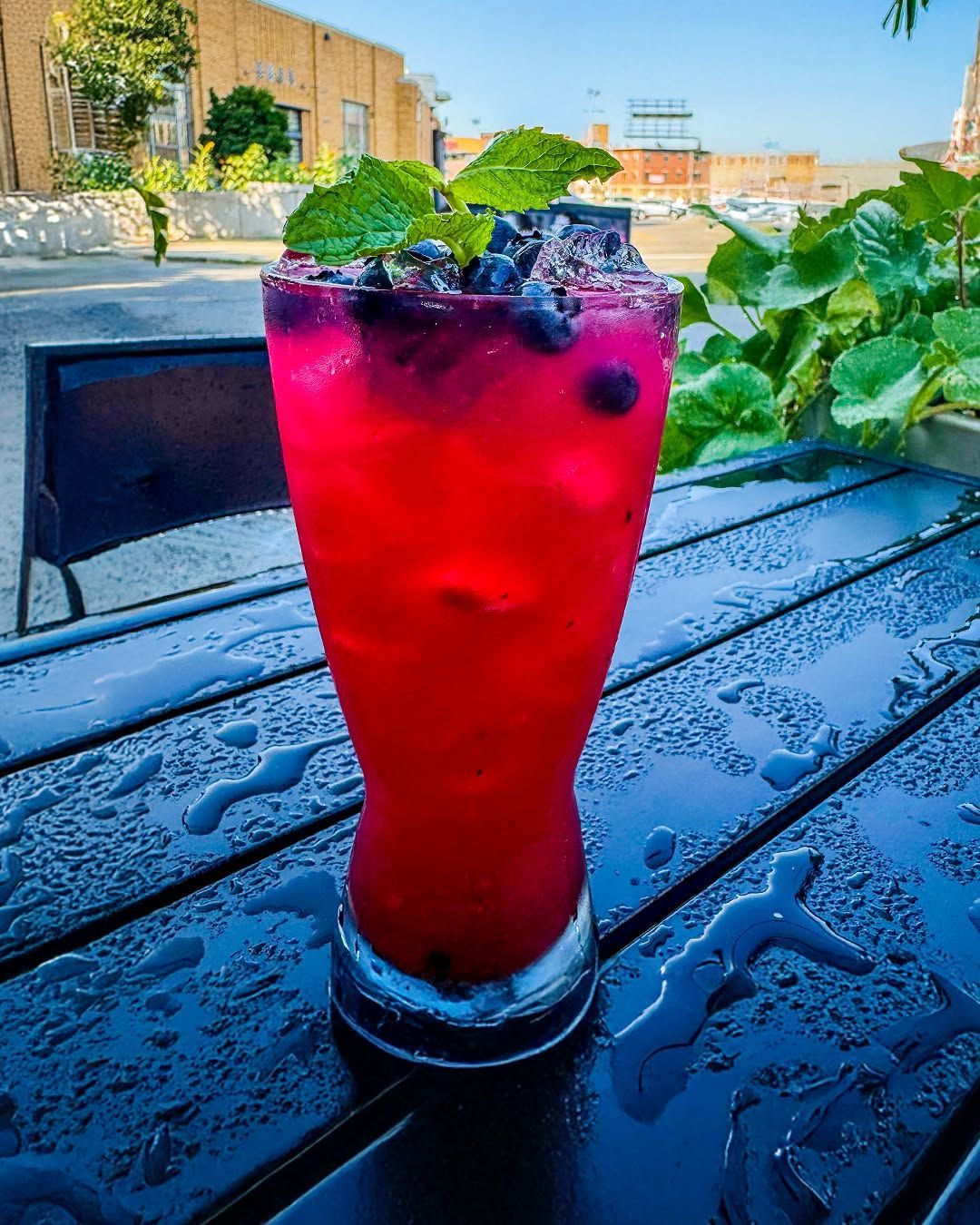 A vibrant red cocktail with blueberries and mint garnish, sitting on a wet, blue table. Outdoor setting.