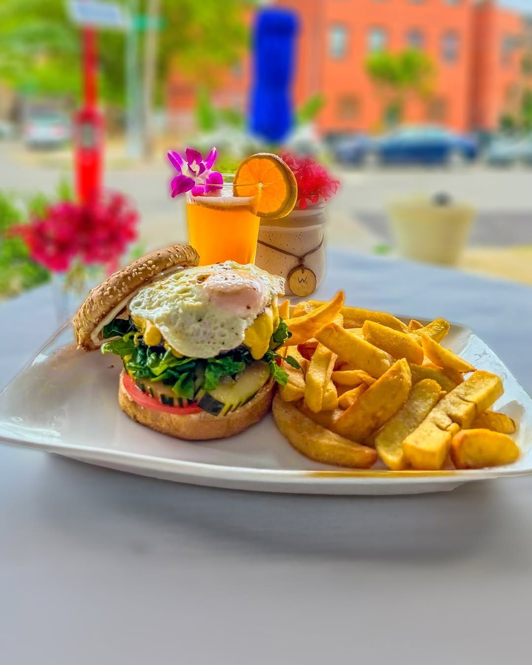 Burger with a fried egg, fries, and orange juice on a white plate, outside a cafe.