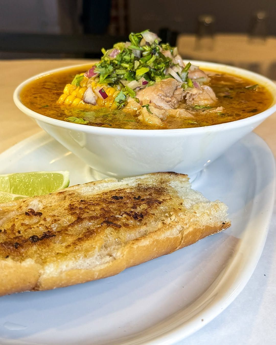 Bowl of soup with corn, meat, and garnish, served with toasted bread and lime.