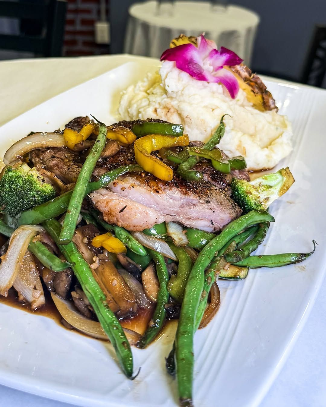 Grilled steak with stir-fried vegetables and mashed potatoes, garnished with a flower, on a white plate.