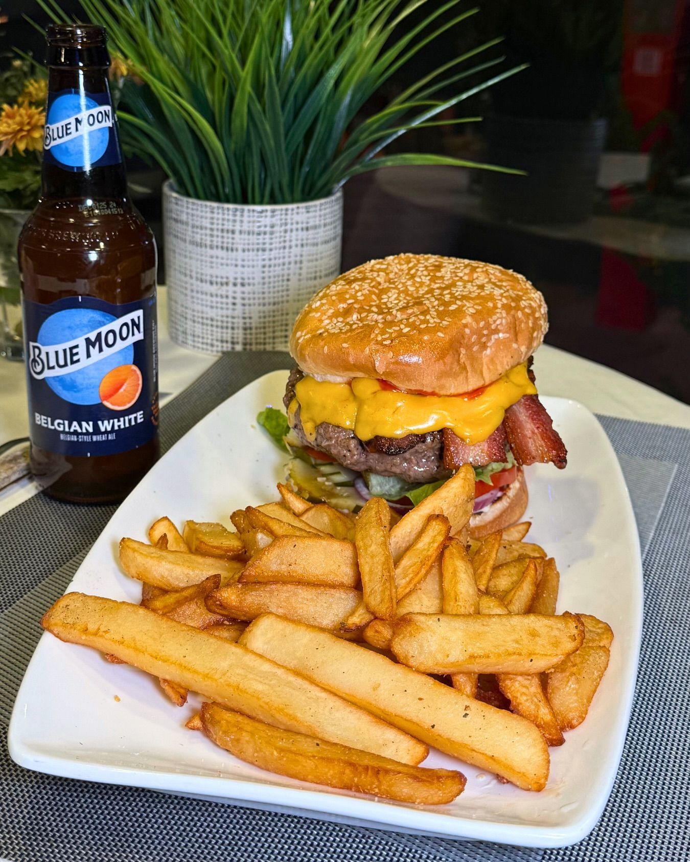 Burger, fries, and Blue Moon beer on a plate. The burger has cheese, bacon, and toppings.