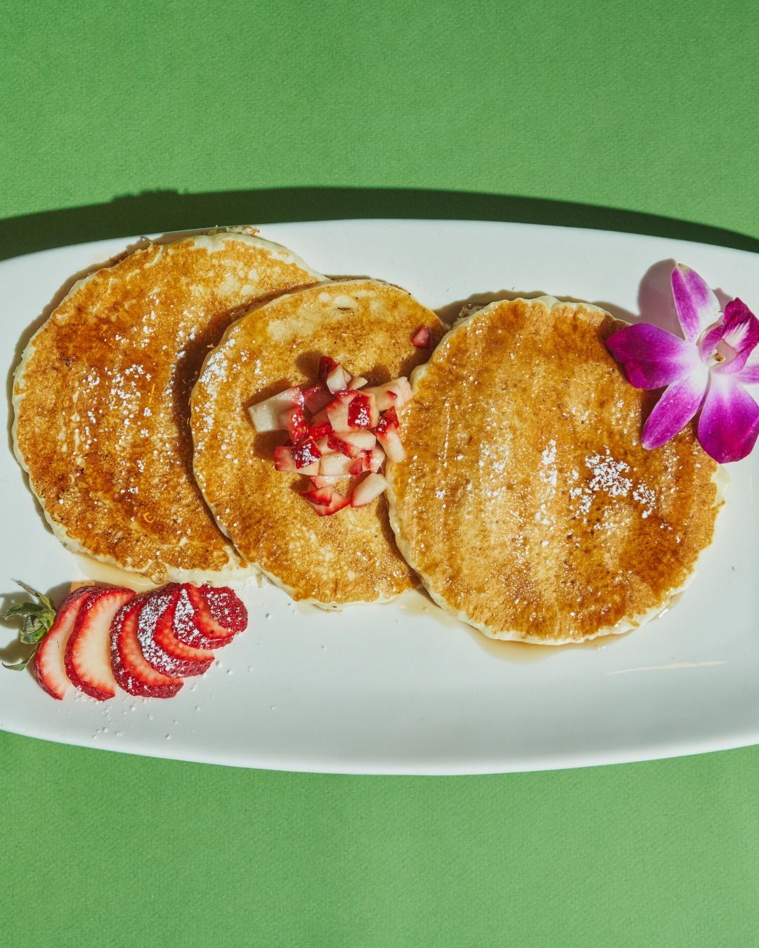 Three golden pancakes on a white plate, topped with fruit and a purple flower, on a green background.