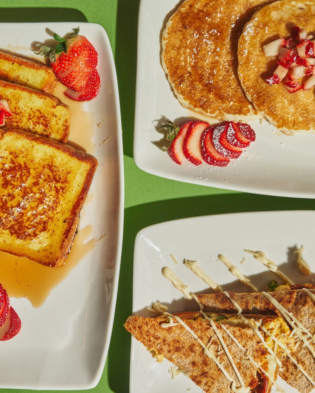 Plates of breakfast food: French toast, pancakes, and a sandwich, with strawberries on a green surface.