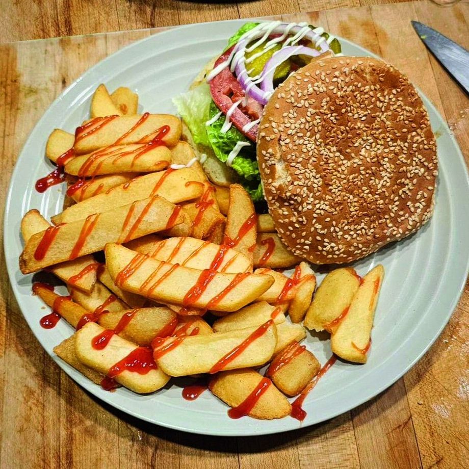 Plate with burger, sesame seed bun, lettuce, pickles, red onion, ketchup-covered potato wedges.