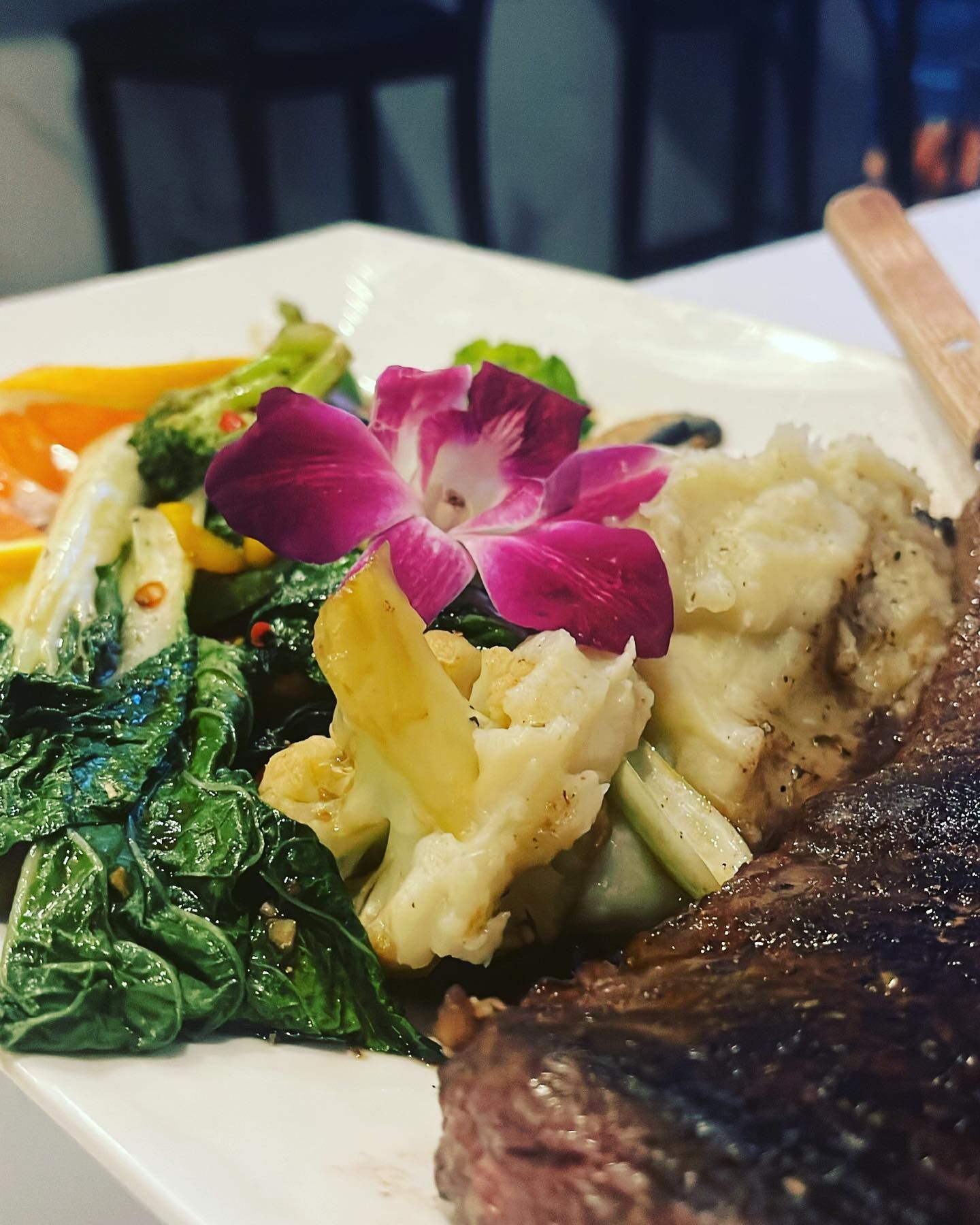 Close-up of grilled steak with mashed potatoes and vegetables, garnished with a purple flower.