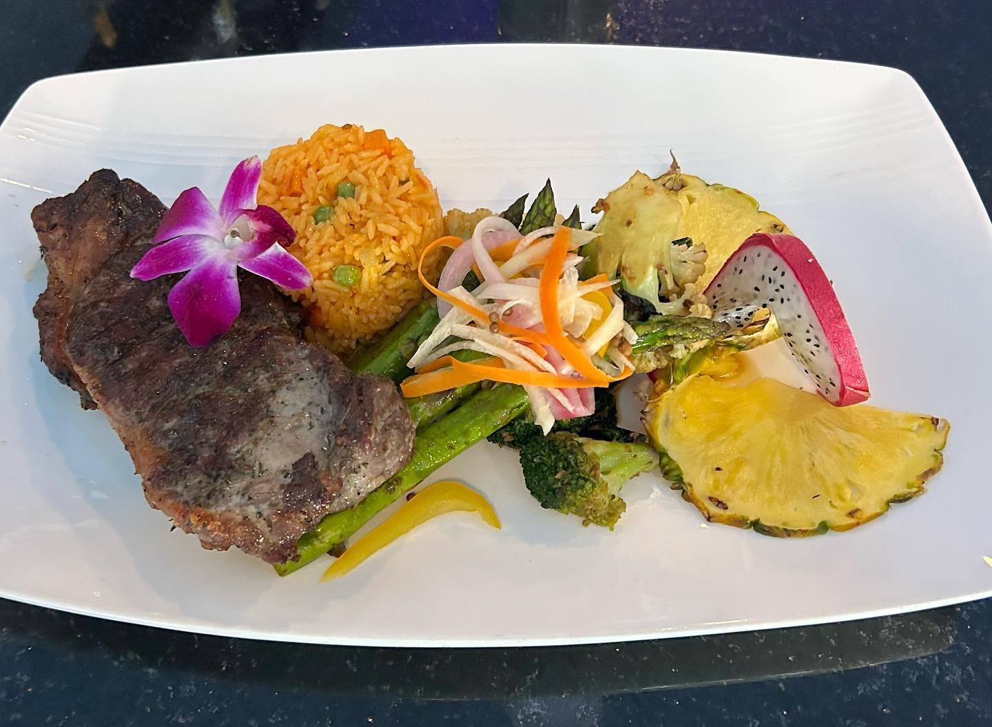 Plate of food: grilled meat, rice, vegetables, pineapple, dragon fruit, and orchid.