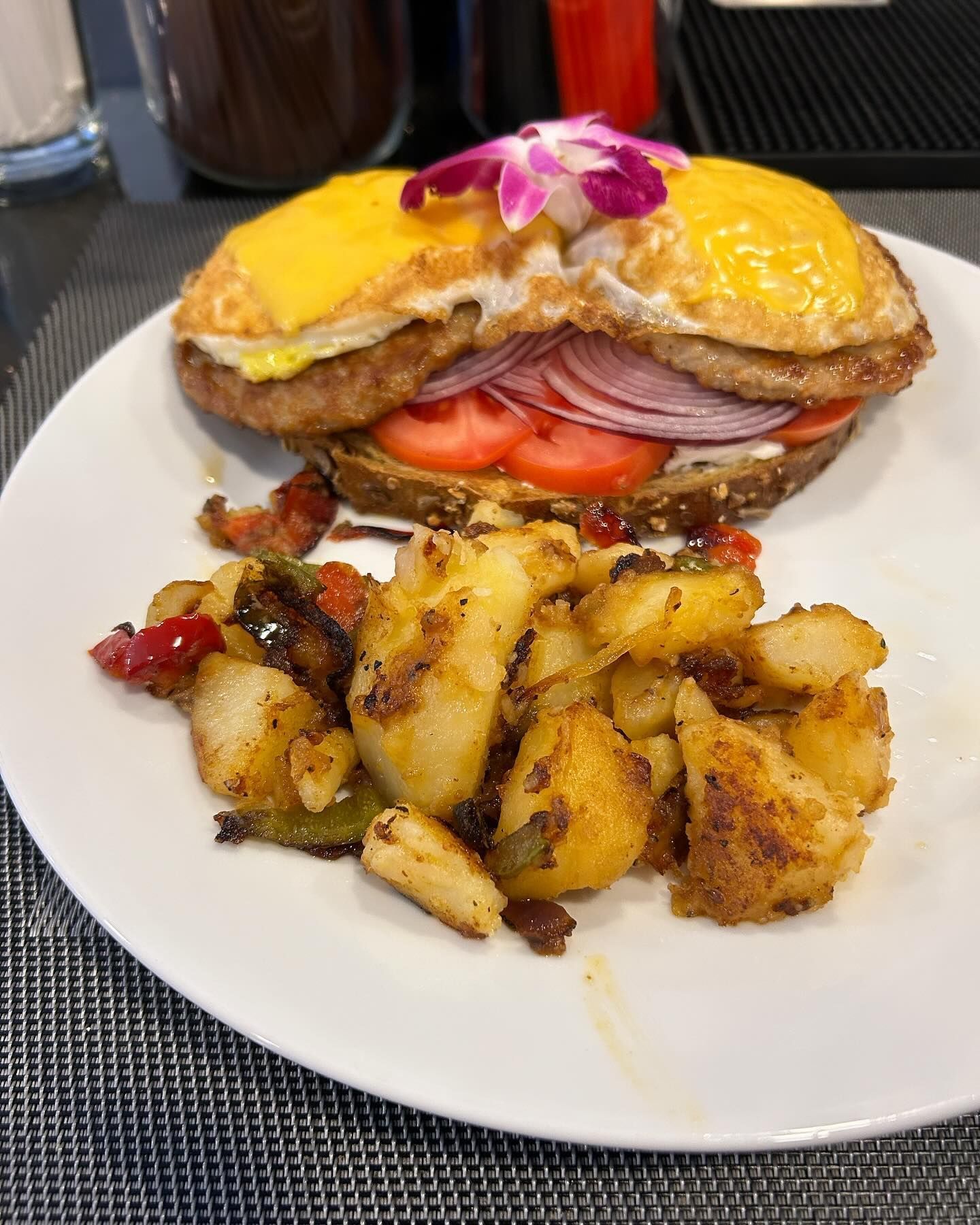 Breakfast sandwich with fried potatoes on a white plate. Sandwich has eggs, tomato, red onion and cheese.
