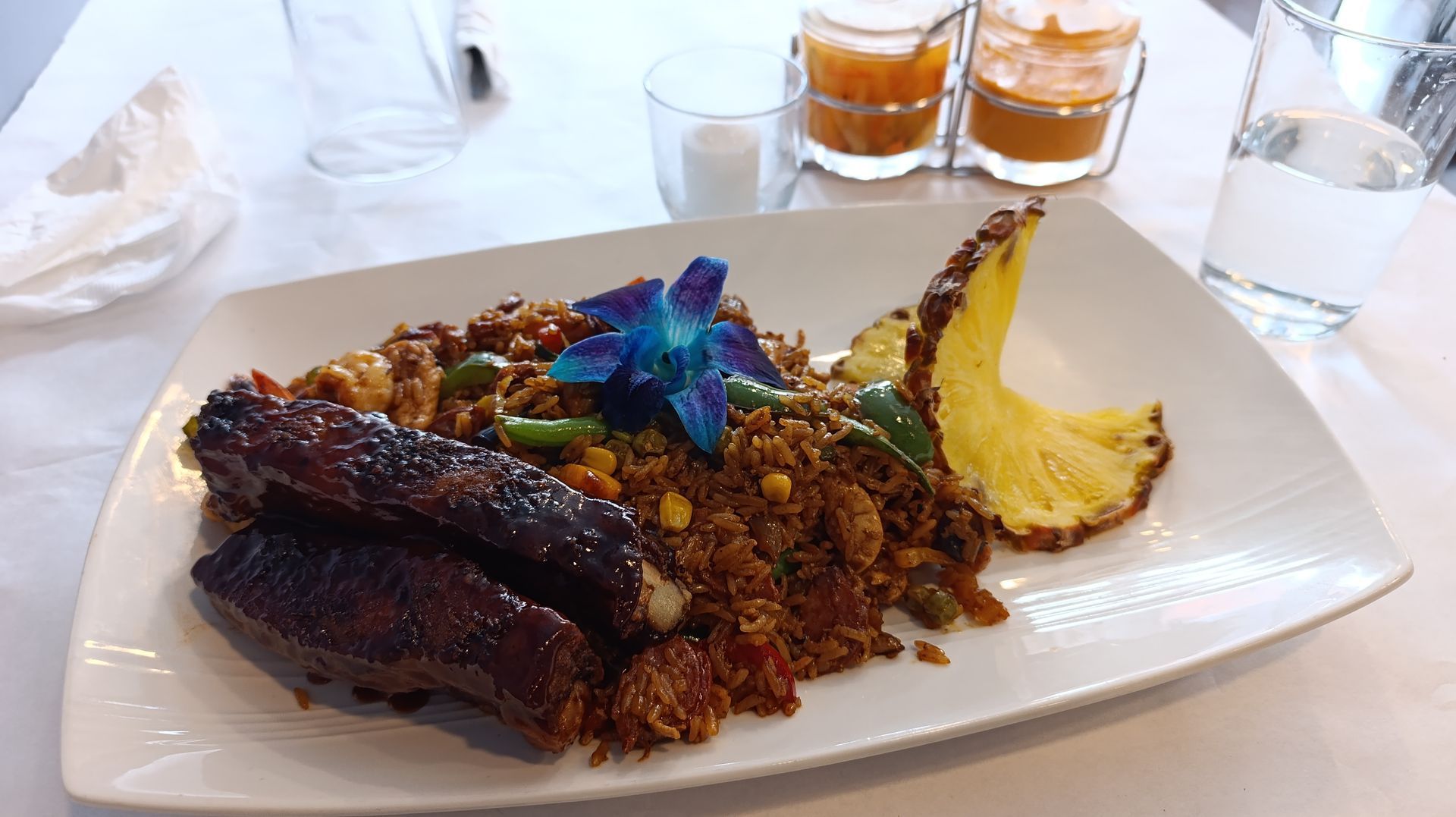 Plate of food: fried rice with sausage and pineapple, garnished with a blue flower. Drinks and water glass.