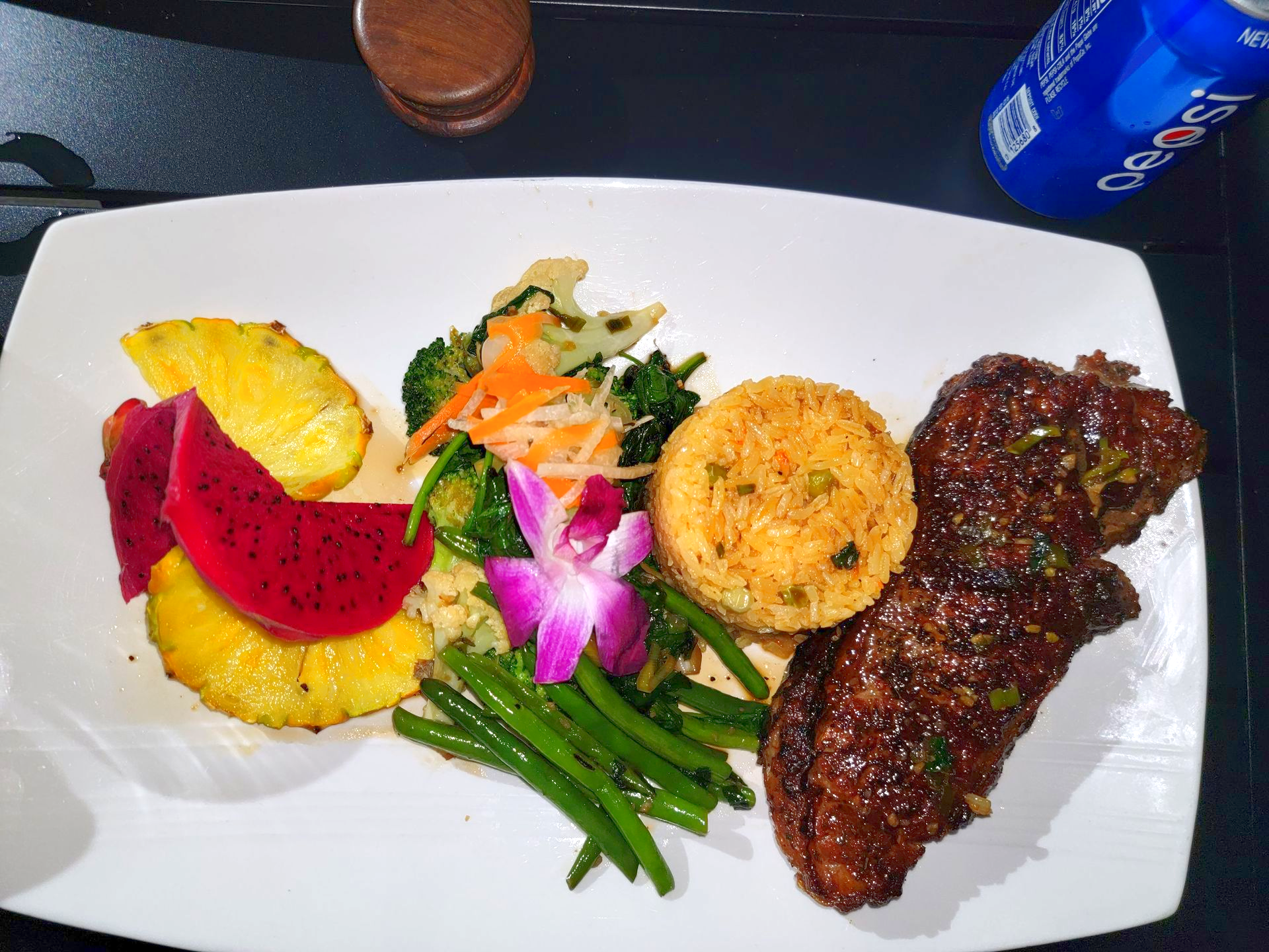 Plate of food with steak, rice, vegetables, and fruit. A Pepsi can is visible.