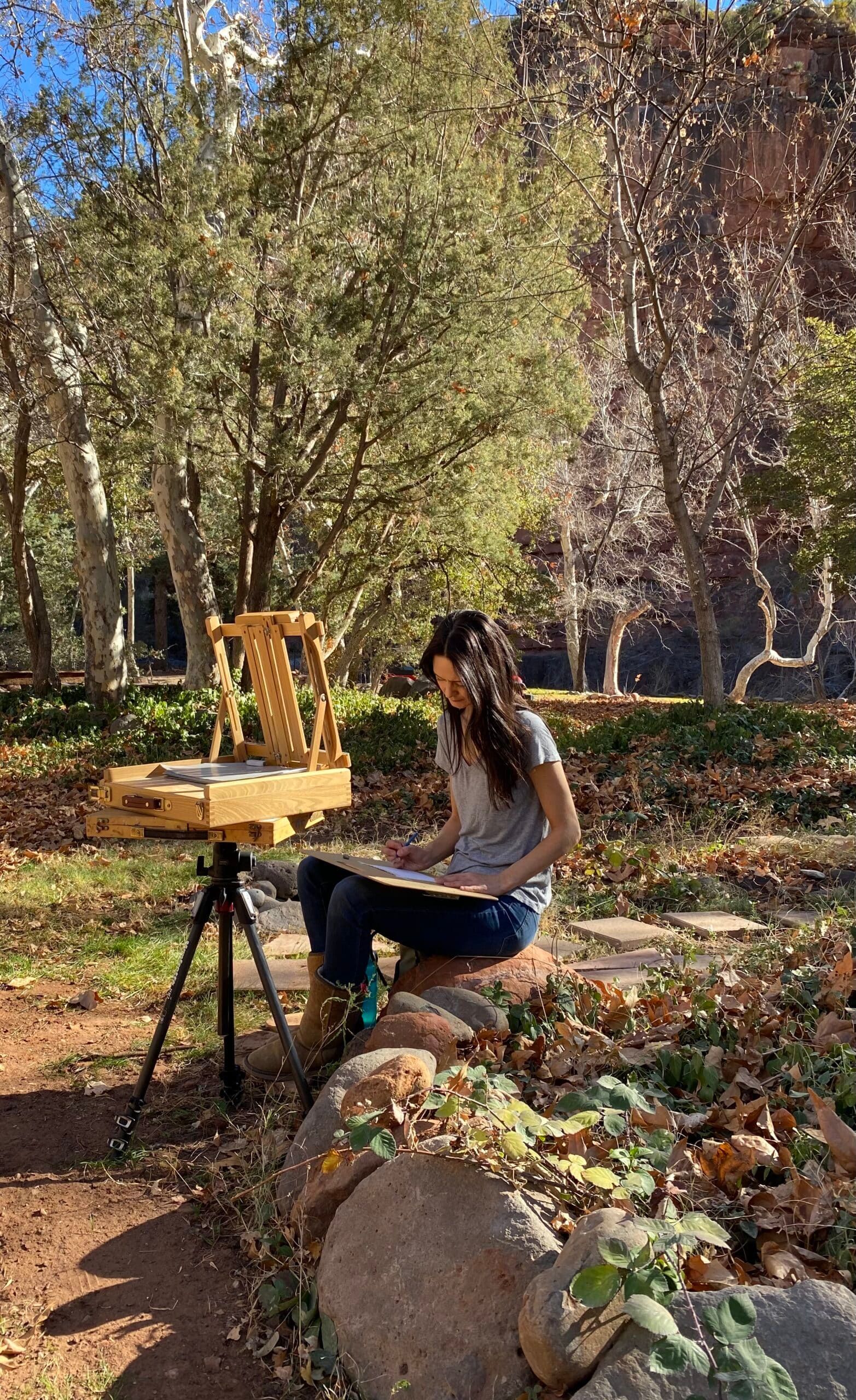 A woman is sitting on a rock drawing in the woods.
