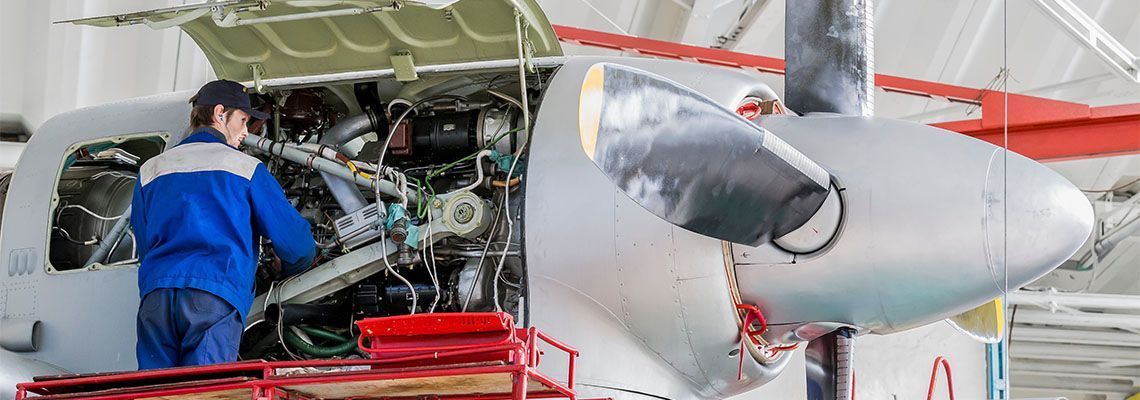 Technician in blue coveralls inspecting a jet engine beside a silver aircraft wing