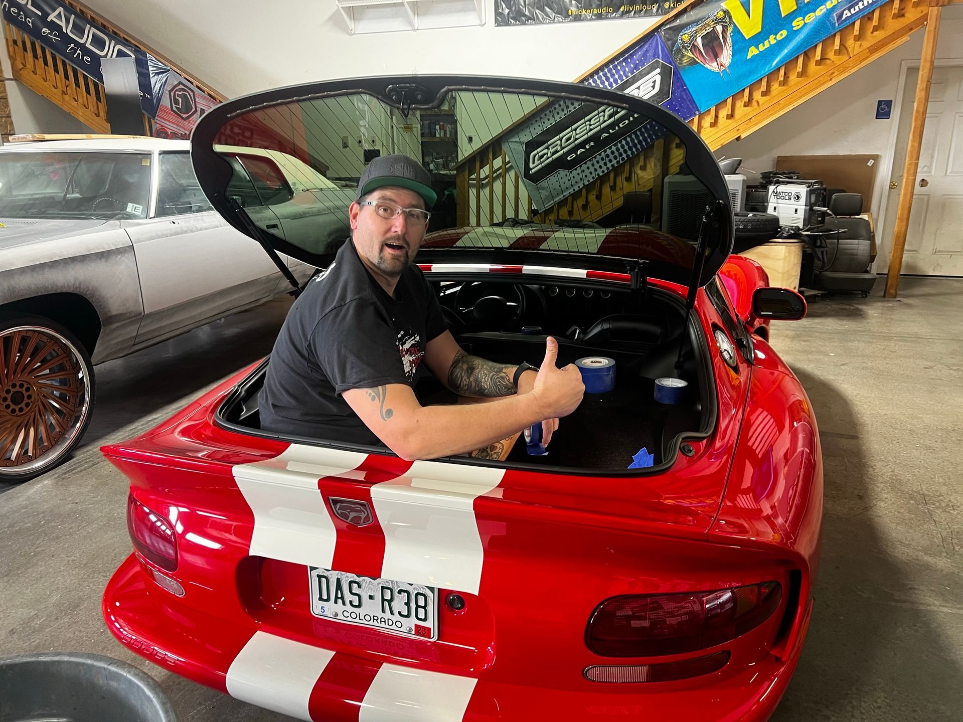 A man is sitting in the back of a red sports car giving a thumbs up.