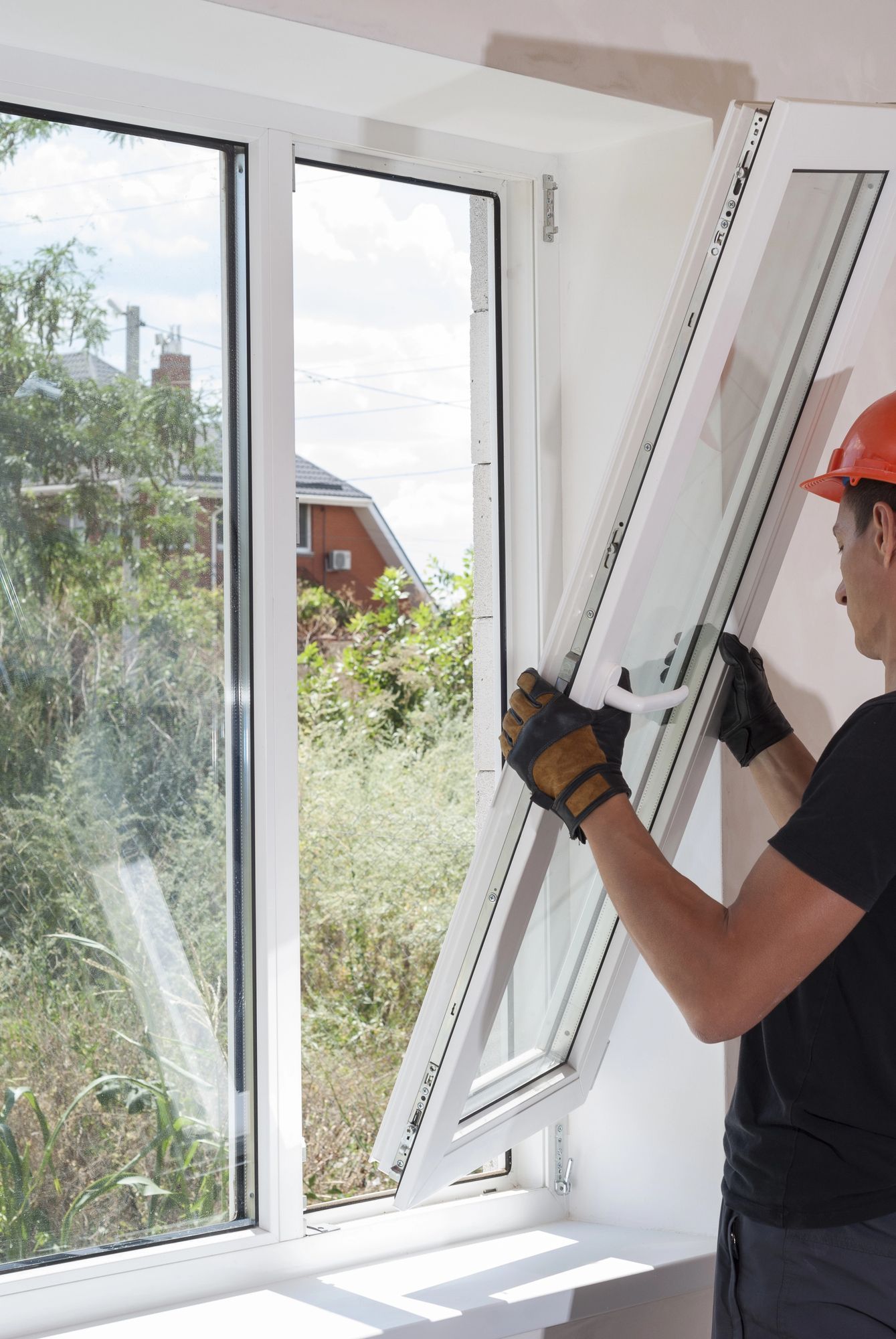 Persona que instala un marco de ventana blanco junto a una ventana abierta, con guantes y casco rojo.