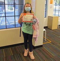 A young girl wearing a mask is holding a certificate in a room.
